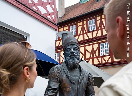 Adam Riese Denkmal (Bad Staffelstein, Obermain.Jura) Zwei Personen betrachten eine Bronzestatue in historischer Kleidung vor Fachwerkhäusern.